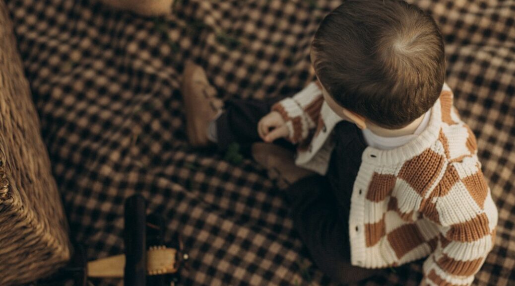 A baby sits on a checkered blanket with a basket